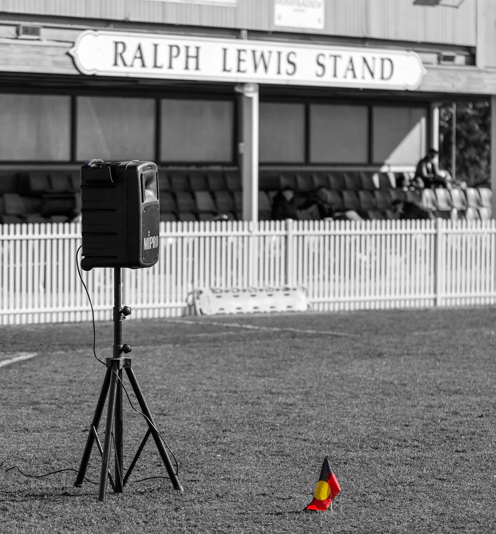 Indigenous Round Flag Grey scale image of Ralph Lewis Stand, a speaker on a football field, and the Aboriginal Flag, The flag retains its normal colours while the rest of the image is greyscale.