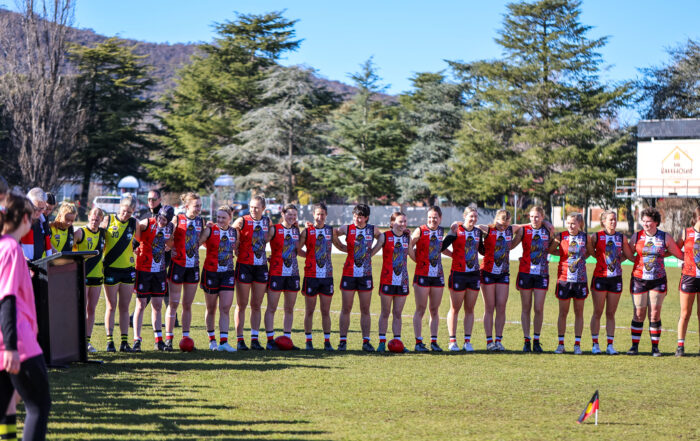 Ainslie Womens team standing shoulder to shoulder infront of a small aboriginal flag planted in the ground