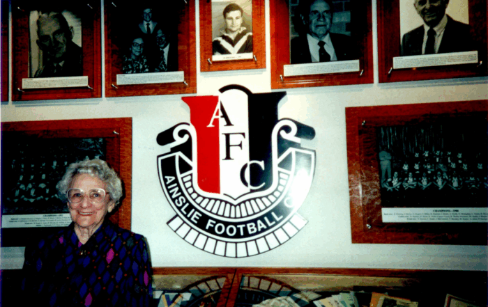 Image of Catharine "Kitty" James standing infront of wall that displays photos of Ainslie players, Ainslie logo.