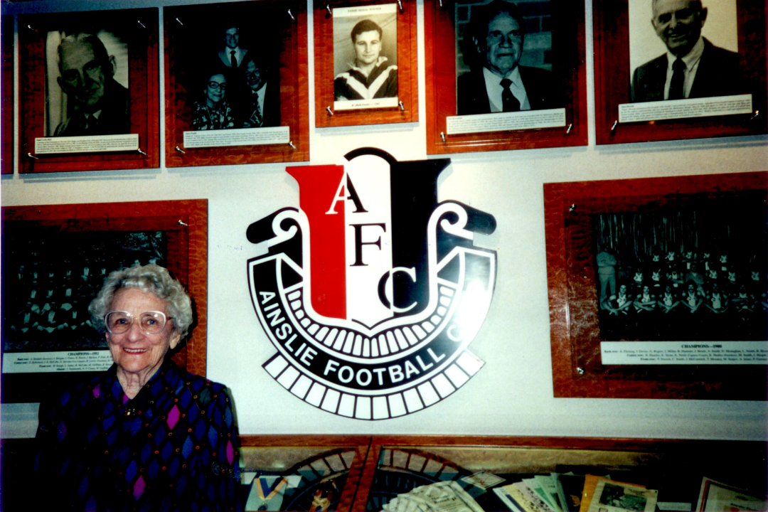 Image of Catharine "Kitty" James standing infront of wall that displays photos of Ainslie players, Ainslie logo.