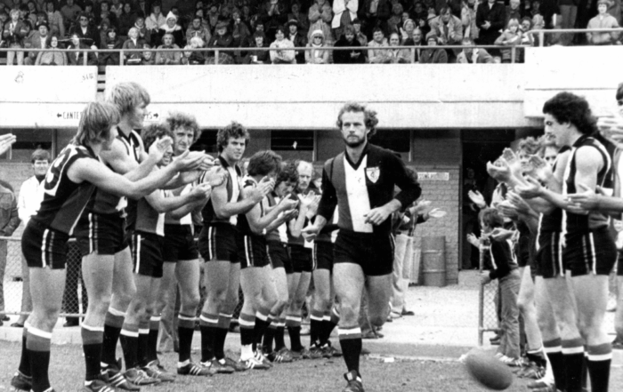 Black and white photo of Charles running onto the field while his team are stood either side of him cheering him on.